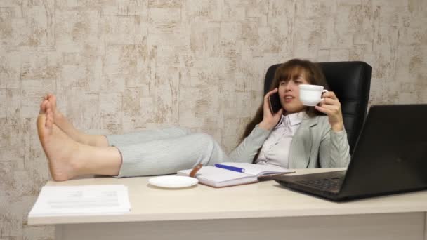 Mooie zakenvrouw zitten in een leunstoel met blote voeten op de tafel rust en drinken thee, koffie. meisje op het werk op kantoor onderhandelt op mobiele telefoon en werkt op een computer. — Stockvideo