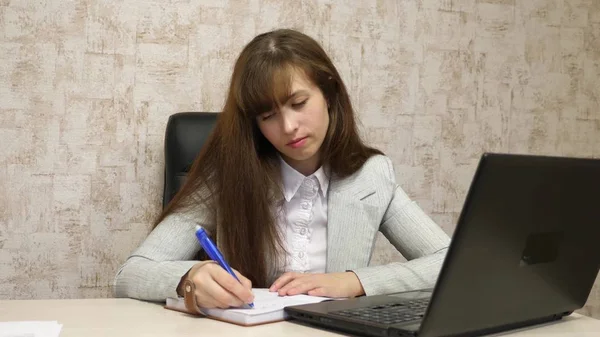 Chica sentada en silla en la oficina en el ordenador y toma notas en el cuaderno. joven empresaria que trabaja detrás de la computadora portátil. secretario escribe pluma en un cuaderno —  Fotos de Stock
