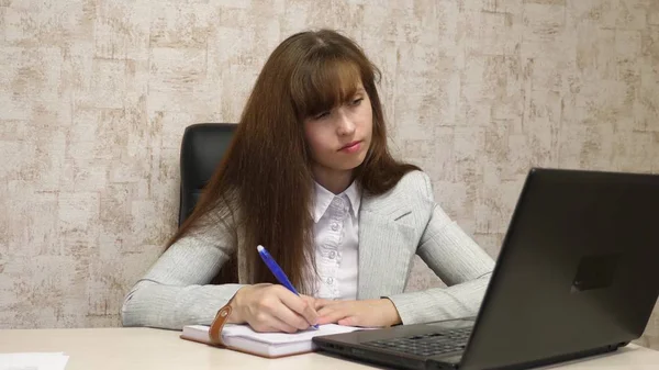 Chica sentada en silla en la oficina en el ordenador y toma notas en el cuaderno. joven empresaria que trabaja detrás de la computadora portátil. secretario escribe pluma en un cuaderno — Foto de Stock