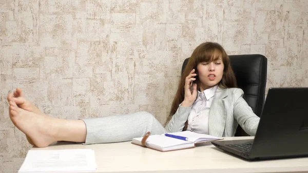 Biznes kobieta siedzi w fotelu z bosymi stopami na stole. Dziewczyna w pracy w biurze na telefon na stole i odpoczynku. — Zdjęcie stockowe