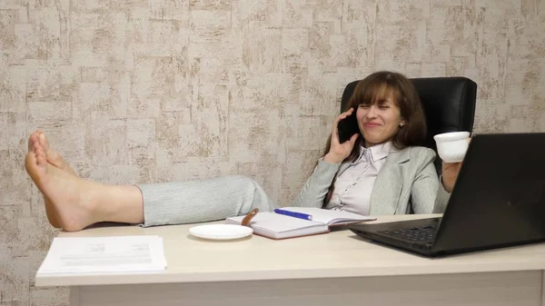 Belle femme d'affaires assise dans un fauteuil avec pieds nus sur la table reposant et buvant du thé, du café. fille au travail au bureau négocie sur téléphone mobile et travaille sur un ordinateur . — Photo