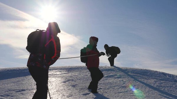 three Alpenists climb rope on snowy mountain. Tourists work together as team shaking heights overcoming difficulties. silhouettes of travelers rise to their victory up hill on ice in rays of the sun.