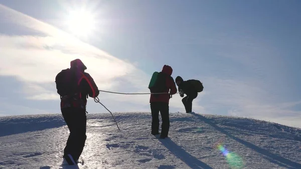 stock image three Alpenists climb rope on snowy mountain. Tourists work together as team shaking heights overcoming difficulties. silhouettes of travelers rise to their victory up hill on ice in rays of the sun.