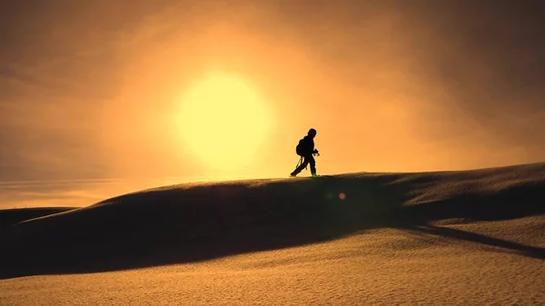 Fotograf resenär i vinter går på snöiga ås i strålar av gula solnedgången. Mountaineer med kamera och stativ promenader i snö längs toppen av en Gustav. — Stockfoto