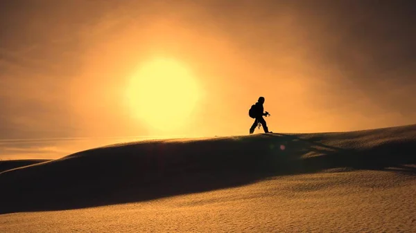 Фотограф-путешественник зимой отправляется на снежный хребет в лучах желтого заката. альпинист с фотоаппаратом и штативом идет по снегу по вершине голома . — стоковое фото