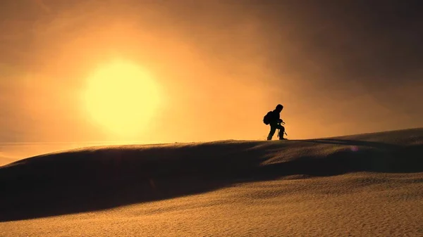 Fotograf resenär i vinter går på snöiga ås i strålar av gula solnedgången. Mountaineer med kamera och stativ promenader i snö längs toppen av en Gustav. — Stockfoto