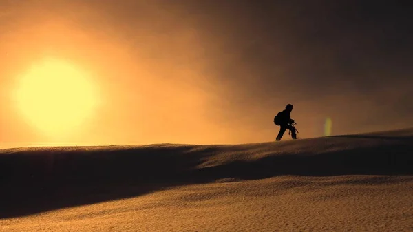 Fotógrafo viajero en invierno va en cresta nevada en los rayos de la puesta del sol amarillo. montañista con cámara y trípode está caminando en la nieve a lo largo de la parte superior de un holom . — Foto de Stock