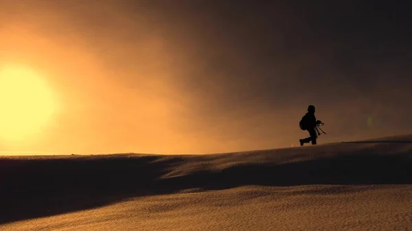 Fotógrafo viajero en invierno va en cresta nevada en los rayos de la puesta del sol amarillo. montañista con cámara y trípode está caminando en la nieve a lo largo de la parte superior de un holom . — Foto de Stock