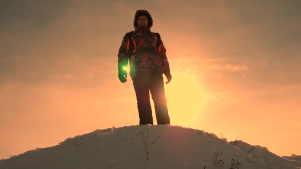Turista se encuentra en la cima de la colina nevada en los rayos del sol brillante. El viajero desciende de las montañas sobre un fondo de puesta de sol amarilla. el trabajo de las personas en condiciones difíciles . — Vídeos de Stock