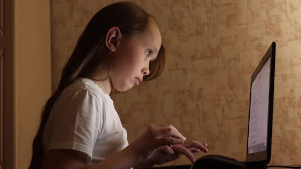 child plays on computer in evening in the room. young girl does her homework on laptop. girl types in search query on a laptop.