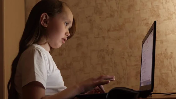 Dítě hraje na počítači večer v místnosti. mladá dívka dělá její domácí úkoly na notebooku. dívka typy v vyhledávací dotaz na notebooku. — Stock fotografie