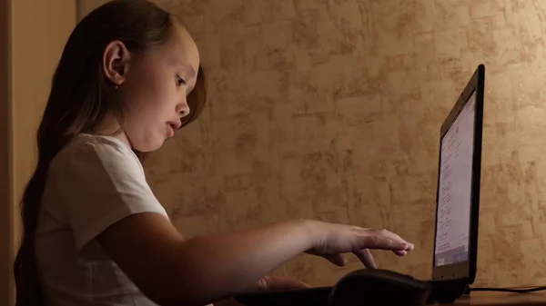 Kind speelt op de computer in de avond in de kamer. jonge meisje doet haar huiswerk op laptop. meisje typen in een zoekopdracht op een laptop. — Stockfoto