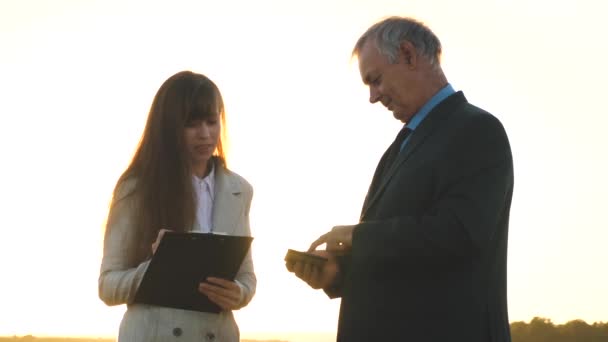 Teamwork men and women on a business project. Businessman and businesswoman with tablet negotiate at sunset and laugh. Business meeting men and women in the park. — Stock Video