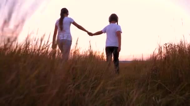 Šťastné děti pěšky přes pole, drželi se za ruce. koncept šťastné dětství. děti ve sluchátkách poslouchat hudbu a hrát na hřišti při západu slunce. — Stock video