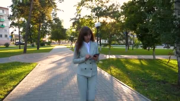 Une fille sensuelle en costume d'affaires léger se rend au travail. belle femme d'affaires marchant le long de l'allée dans le parc avec une tablette dans ses mains . — Video