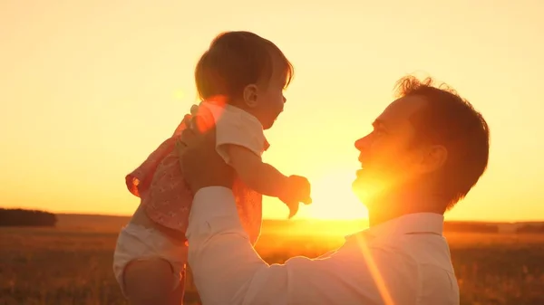 A bela filha em mãos do pai sorri ao pôr-do-sol brilhante. Pai feliz brincando com o bebê no parque em uma noite de verão . — Fotografia de Stock