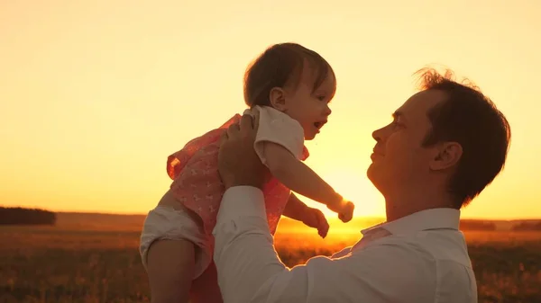 Mooie dochter in handen van de vader glimlach bij heldere zonsondergang. Gelukkig Papa spelen met baby in park op een zomeravond. — Stockfoto