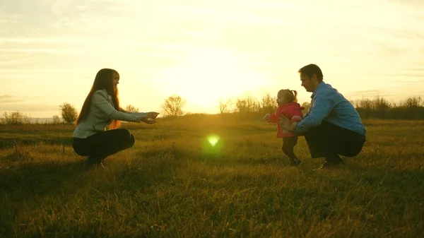 Mama i tata grać z dzieckiem na trawie o zachodzie słońca. koncepcja szczęście rodzinne. dziecko idzie na trawnik od taty do mamy. dziecko bierze pierwsze kroki w parku. — Zdjęcie stockowe