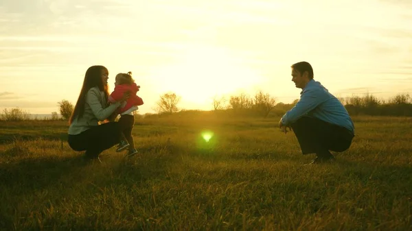 Mama und Papa spielen mit Kindern auf dem Rasen bei Sonnenuntergang. Familienglück. Baby geht auf Rasen von Papa zu Mama. Kind macht erste Schritte im Park. — Stockfoto