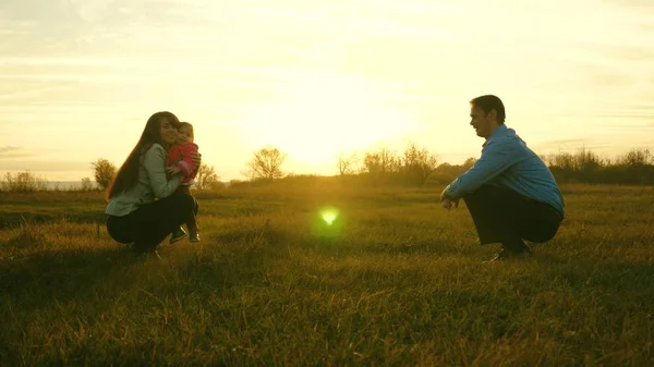 Mama und Papa spielen mit Kindern auf dem Rasen bei Sonnenuntergang. Familienglück. Baby geht auf Rasen von Papa zu Mama. Kind macht erste Schritte im Park. — Stockfoto