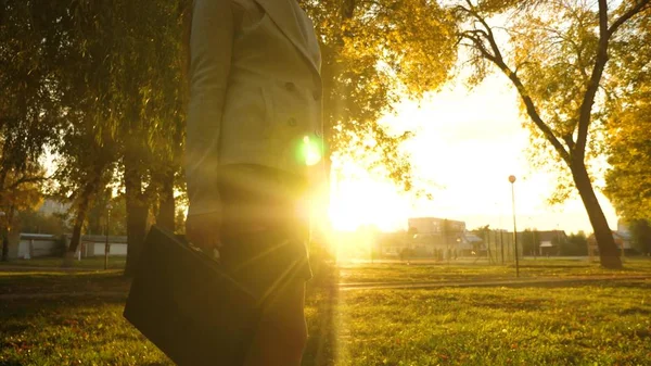 Mujer lleva maletín en la mano y camina por la acera en el parque en los rayos del sol brillante. primer plano . —  Fotos de Stock