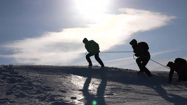 チームワークを獲得する欲望。ロープの上の登山者は、丘の頂上に登る友人を助けます。冬の太陽の明るい光の丘の上の方のシルエット。スポーツ観光の概念. — ストック写真