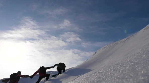 Desejo de trabalho em equipe para ganhar. Escaladores derajat uns aos outros mãos para ajudar um amigo a subir ao topo de uma montanha nevada. viajantes no inverno em uma colina em raios brilhantes do sol. conceito de turismo desportivo . — Fotografia de Stock