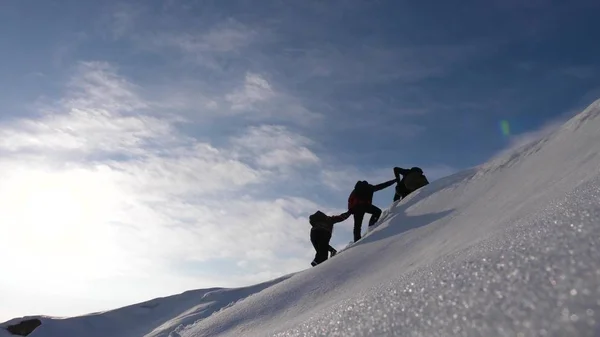 Teamwork will siegen. Bergsteiger entgleisen einander die Hände, um einem Freund beim Aufstieg auf einen schneebedeckten Berg zu helfen. Reisende im Winter auf einem Hügel in hellen Sonnenstrahlen. Konzept des Sporttourismus. — Stockfoto