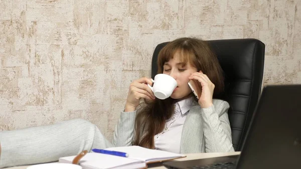 Mulher no escritório bebendo café descansando no local de trabalho. bela empresária com telefone senta-se em uma cadeira com os pés descalços sobre a mesa descansando. menina no trabalho no escritório falando em um telefone celular — Fotografia de Stock