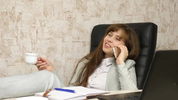 Mujer en la oficina tomando café descansando en el lugar de trabajo. hermosa mujer de negocios con teléfono se sienta en una silla con los pies descalzos en la mesa descansando. chica en el trabajo en la oficina hablando en un teléfono móvil —  Fotos de Stock