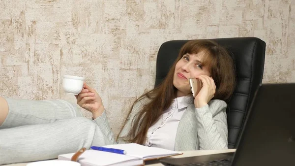 Mujer en la oficina tomando café descansando en el lugar de trabajo. hermosa mujer de negocios con teléfono se sienta en una silla con los pies descalzos en la mesa descansando. chica en el trabajo en la oficina hablando en un teléfono móvil — Foto de Stock