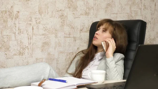 Femme au bureau boire du café reposant sur le lieu de travail. belle femme d'affaires avec téléphone se trouve dans une chaise avec pieds nus sur la table de repos. fille au travail dans le bureau parler sur un téléphone mobile — Photo