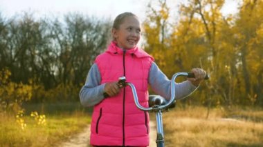 genç kız yolda gidiyor ve bisiklet alır. Çocuk parkta sonbaharda yürür. mutlu kız kameraya sallayarak. Çocuk spor doğa kavramı.