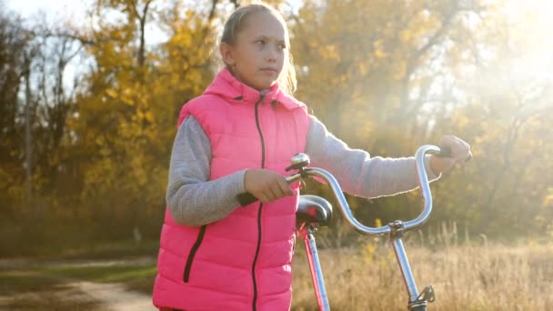 A child with a Bicycle walks in the Park in the autumn in the warm sun. A young girl is on the road and rolls the bike. slow motion. The concept of childrens sports in nature. — Stock Video