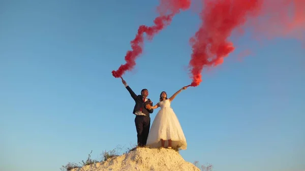 Šťastná nevěsta a ženich mává barevné růžový kouř proti modré obloze a směje se. Líbánky. Romantiku. Vztah mezi mužem a ženou — Stock fotografie