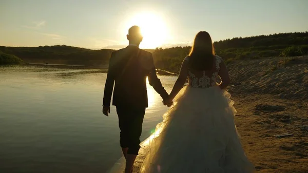 loving couple walks hand in hand on the summer beach in the rays of sunset. The bride in a white dress and the groom barefoot walking on the water on the river bank. Slow motion.