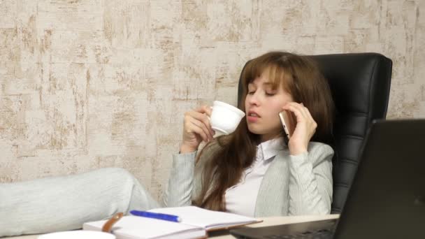 Meisje op het werk in het kantoor te praten op smartphone met kop koffie in de hand. computerwerk. mooie zakelijke vrouw zittend in een stoel met blote voeten op tafel ontspannen en drinken van thee, koffie. — Stockvideo