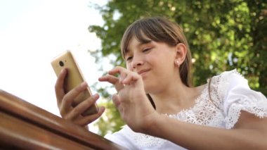 bir parkta bir bankta Internet sayfaları aracılığıyla saygısız smartphone kullanan mutlu kız. yakın çekim. Bin yıllık kadında genç jestleri ekranın üzerinde yapma Botanik Bahçesi,.