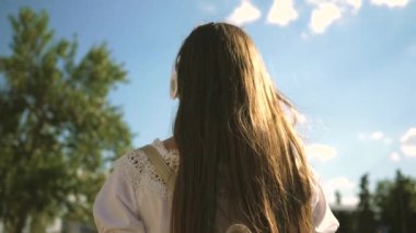 Bir kız kulaklıklar ile bir şehir Sokağı boyunca yürür ve mavi gökyüzüne karşı müzik dinler. şehirde genç kız gezi. uzun saçlı beyaz bir elbise şehir etrafında seyahat kız. Yavaş hareket.