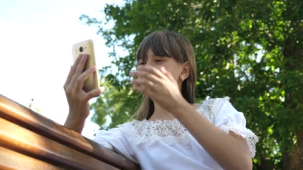 스마트 폰을 사용 하는 어린 소녀가 아름 다운 녹색 공원에 있는 벤치에 편지를 쓰고 있습니다. 수목원에서 젊은 천년 여성, 전화 디스플레이에 제스처 만들기. — 비디오