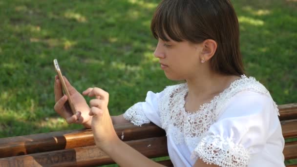 Vacker flicka med en smartphone är att skriva ett brev på en bänk i en vacker grön park. Slow motion. Ung tusenåriga kvinna i Arboretum, göra gester på telefonens display. — Stockvideo