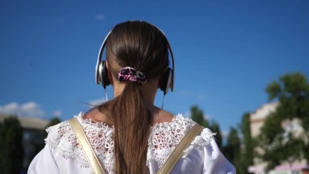 En ung jente går ned en gate med hodetelefoner og hører på musikk. Tenåringsjente i hvit kjole med langt hår reiser rundt i byen mot den blå himmelen. Langsom bevegelse – stockvideo