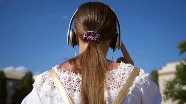 Tenåringsjente i hvit kjole med langt hår reiser rundt i byen mot den blå himmelen. Langsom bevegelse. En ung jente går ned en gate med hodetelefoner og lytter til musikk . – stockvideo