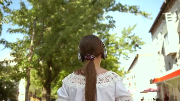 快乐的女孩带着耳机走在城市街道上听音乐。穿着长头发的白色衣服的女孩在城市里四处走动。慢动作。十几岁的女孩在城市郊游. — 图库视频影像