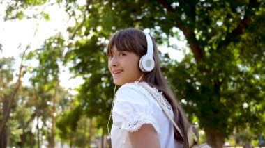 bir sırt çantası ile kız kulaklıklar Park gider ve müzik dinler ve gülümsüyor, Genç mutlu kamera objektifi elini dalgaları. uzun saçlı beyaz bir elbise içinde kız şehir etrafında yolculuk. Yavaş hareket.