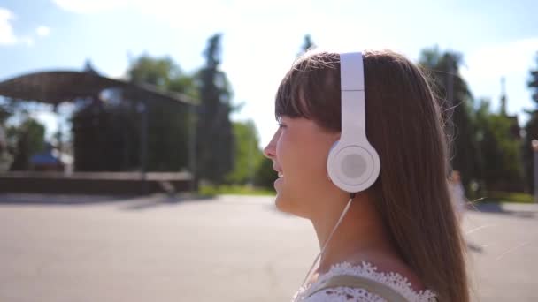 En flicka går längs en gata med hörlurar och lyssnar på musik och leenden. flicka i en vit klänning med långt hår reser runt staden. Slow motion. tonåring flicka utflykt i staden. — Stockvideo