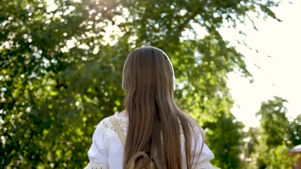 Ein Mädchen mit Rucksack geht mit Kopfhörern in den Park, hört Musik und lächelt winkend mit der Hand und winkt mit sich selbst. Mädchen im weißen Kleid mit langen Haaren reist durch die Stadt. Zeitlupe. — Stockvideo