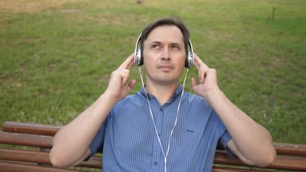 Schöner junger Mann, der sich in einem Park ausruht, Musik von seinem Smartphone mit Kopfhörern hört und draußen in einem Park auf einer Bank in der Mitte Europas tanzt. — Stockvideo