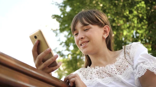 Счастливая девушка с помощью смартфона листает страницы в интернете в парке на скамейке запасных. крупным планом. Молодая женщина тысячелетия в дендрарии, делает жесты на дисплее телефона . — стоковое фото