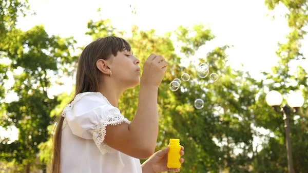 Menina bonita soprando bolhas de sabão no parque na primavera, verão e sorrindo. Movimento lento. jovem brincando no parque . — Fotografia de Stock